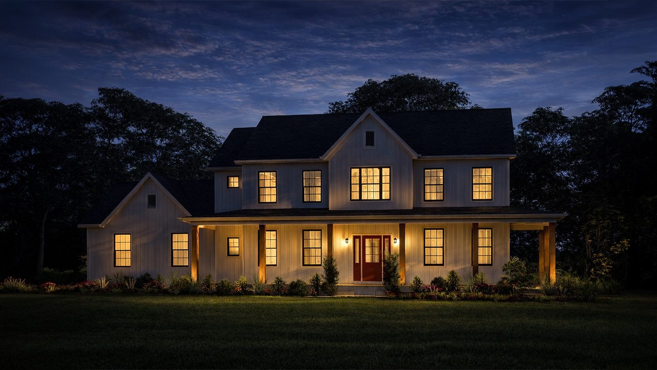 A twilight view of the same two-story modern farmhouse. Warm, yellow light glows prominently from inside all the windows. Exterior sconces illuminate the red front doors, while downlighting from the porch roof highlights the wooden columns and the foundation landscaping. The house stands out brightly against the dark silhouettes of the background trees and a deep blue, dusky sky.