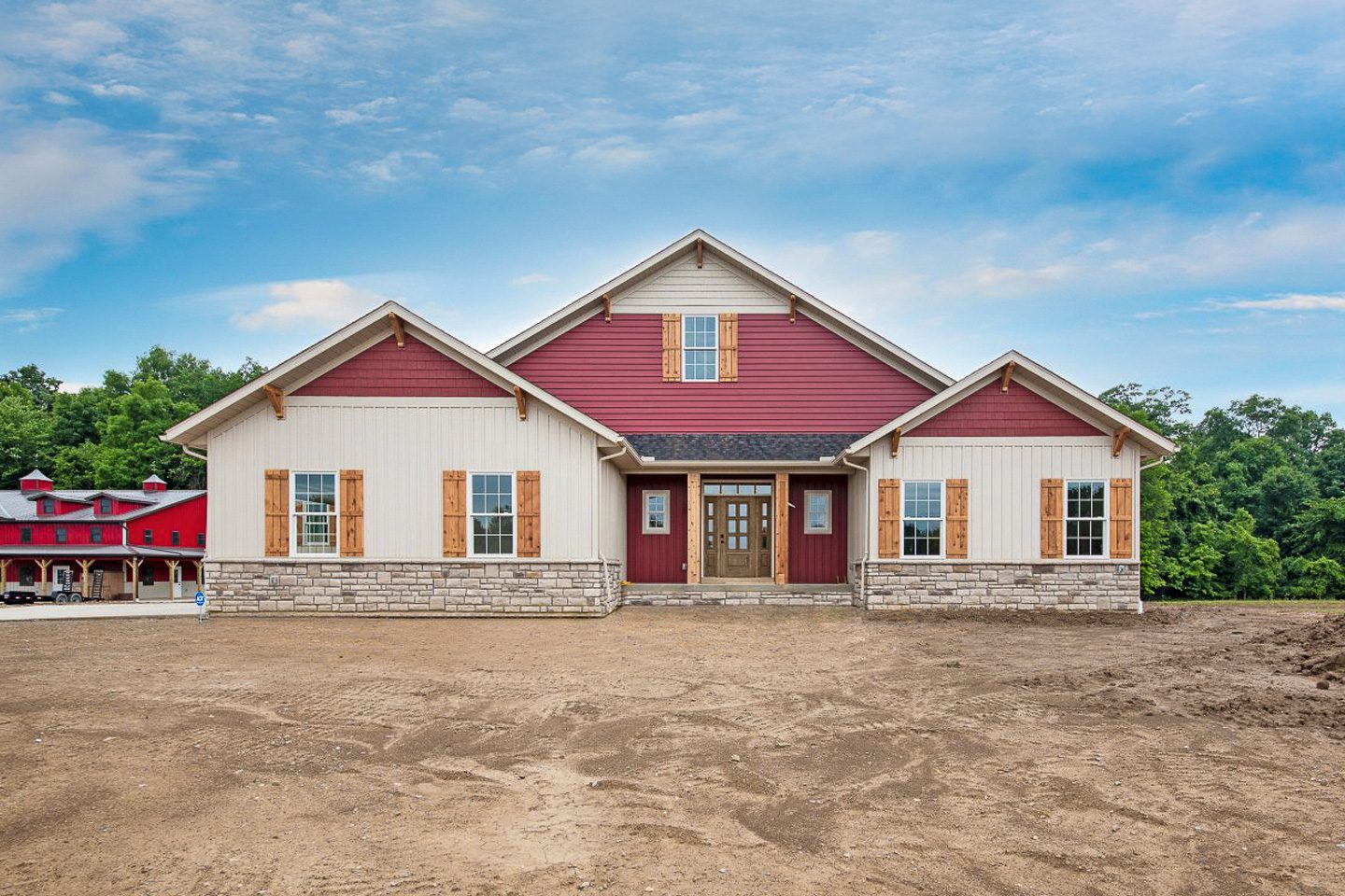 Front exterior of a single-story custom ranch home featuring white siding with red board-and-batten accents, a covered front porch with wood columns, an attached three-car garage, and a spacious homesite under construction.