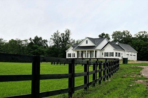 Newman custom Home design with rolling hills and fence. Exterior is white, dark gray shingles and three car garage.