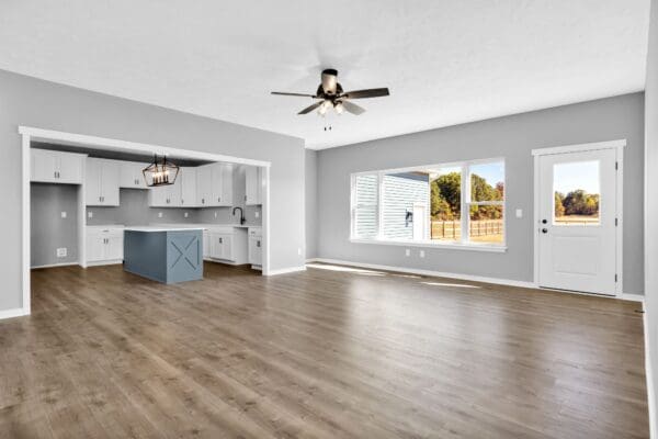 Open-concept great room with vaulted ceilings, light wood flooring, a ceiling fan, large windows overlooking the backyard, and seamless views into the kitchen.